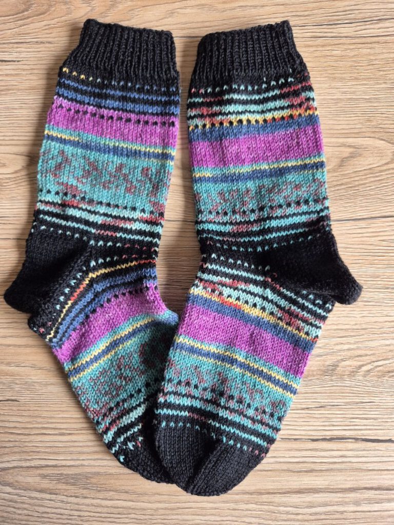 Fertig gestrickte Nebulasocks von @Plutonia42 in schwarz und grau-blau, flach liegend auf einem hellen Holzboden.