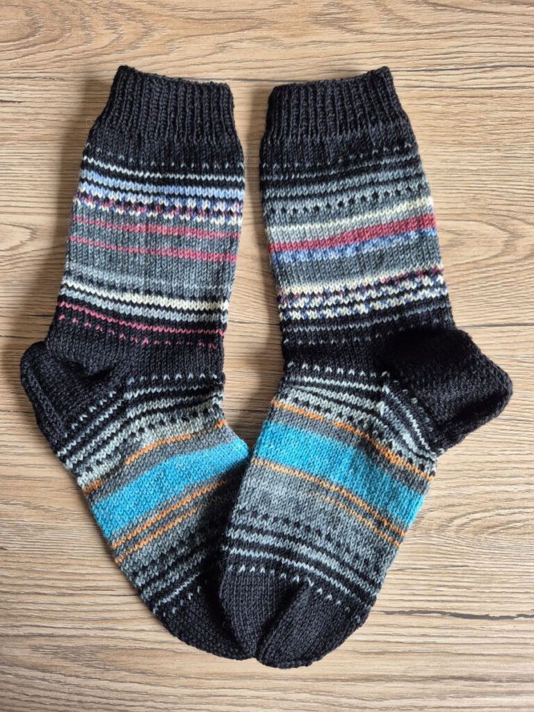 Fertig gestrickte Nebulasocks von @Plutonia42 in schwarz und grau-blau, flach liegend auf einem hellen Holzboden.