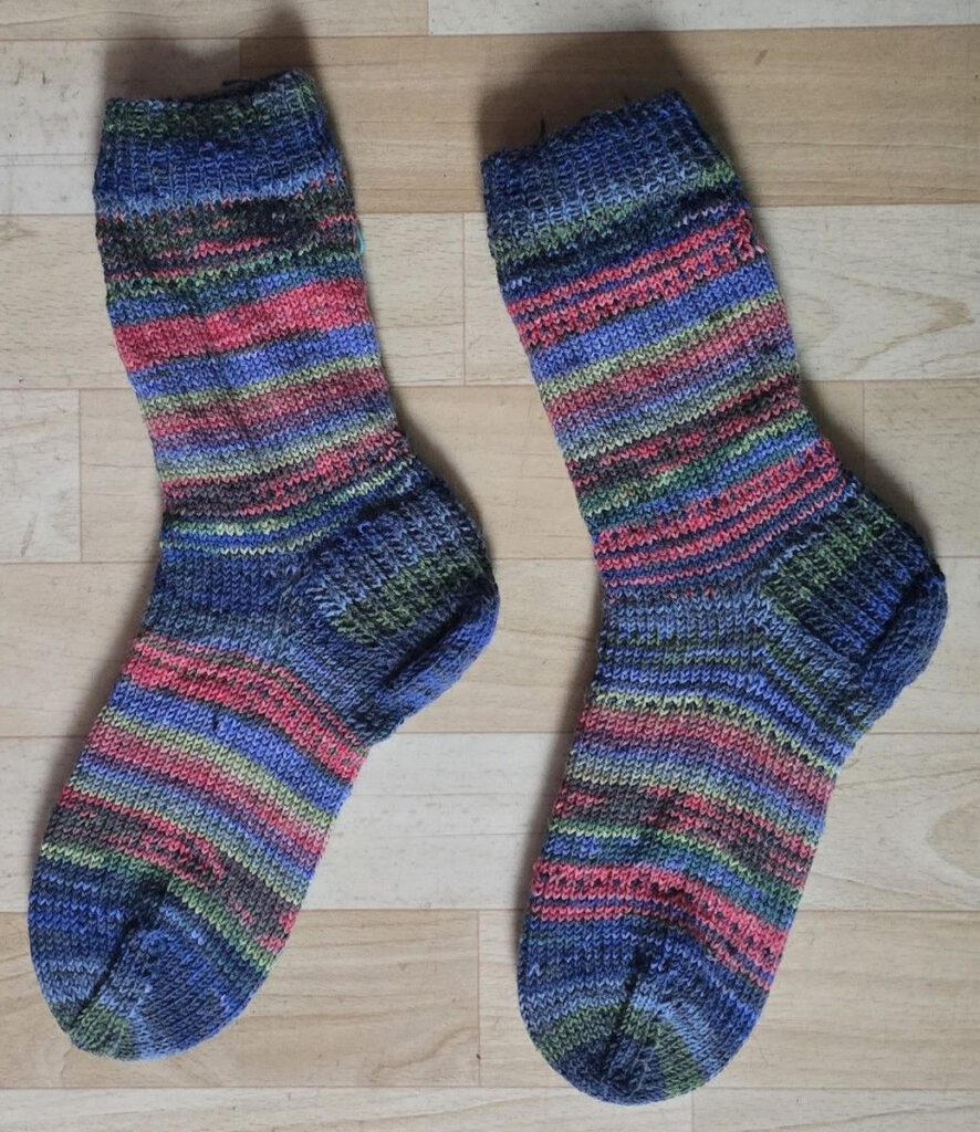 Fertig gestrickte Nebulasocks von @birgittlinnemann in Blau und Pink, flach liegend auf einem hellen Holzboden.