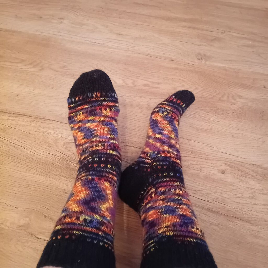 Fertig gestrickte Nebulasocks von Elke in Schwarz und Orange, am Fuß getragen auf einem hellen Holzboden.