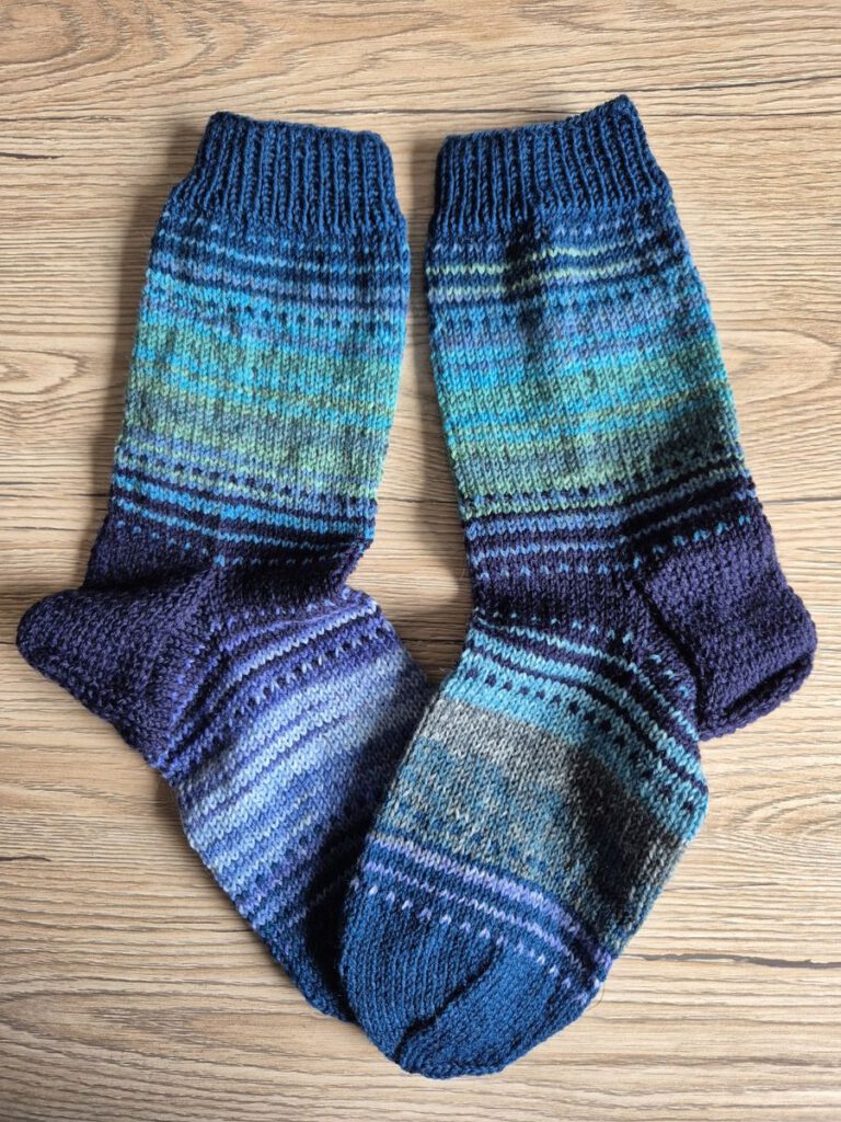Fertig gestrickte Nebulasocks von @Plutonia42 in 5 verschiedenen Farben, flach liegend auf einem hellen Holzboden.