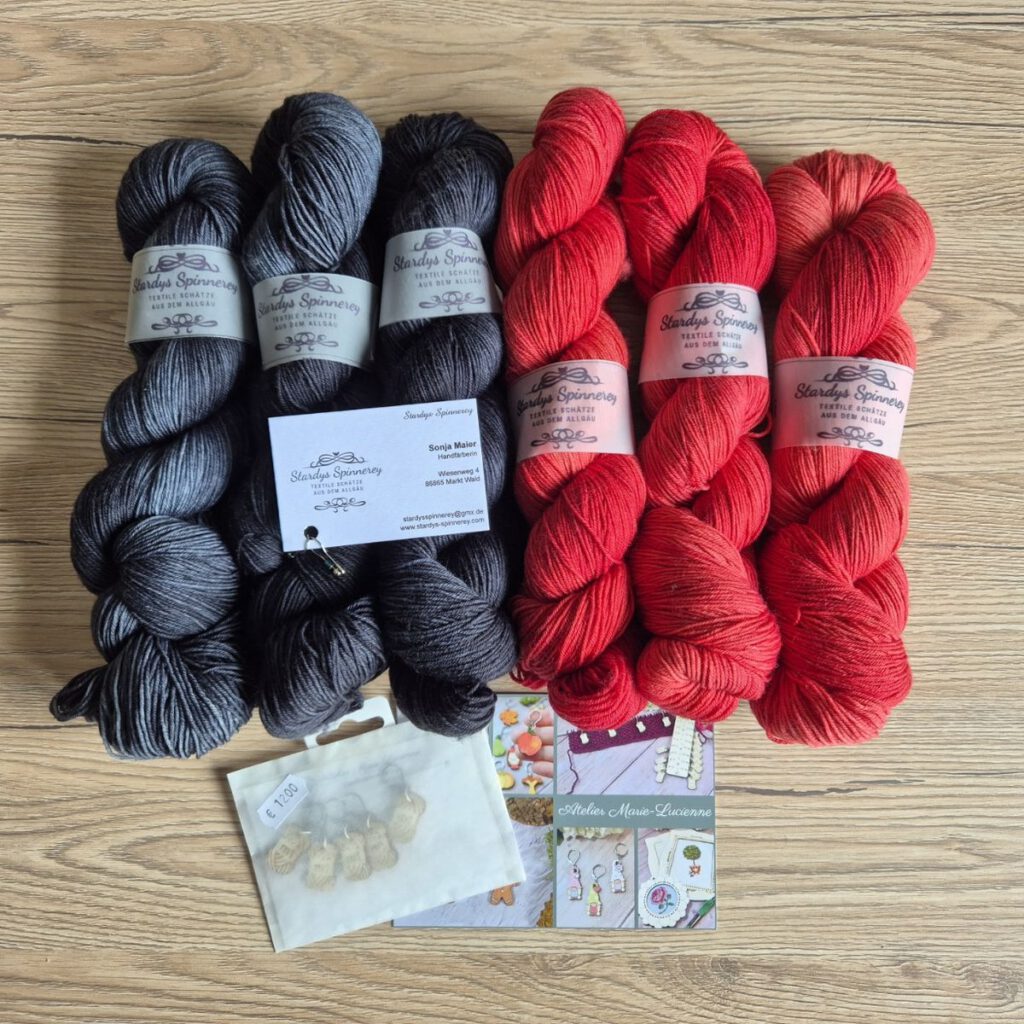 Drei schwarze und drei rote Wollstränge von stardysspinnerey neben dekorativen Maschenmarkierern von ateliermarielucienne auf einem Tisch