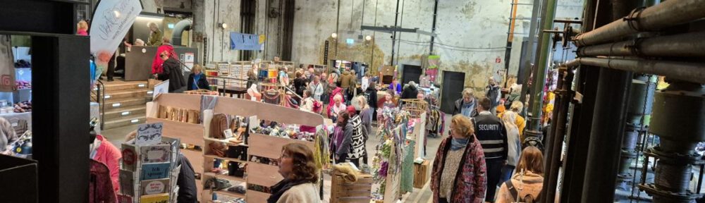Blick in einen gut besuchten, engen Gang einer Messehalle auf dem Wollfest Kielgeholt mit vielen Menschen und bunten Wollständen
