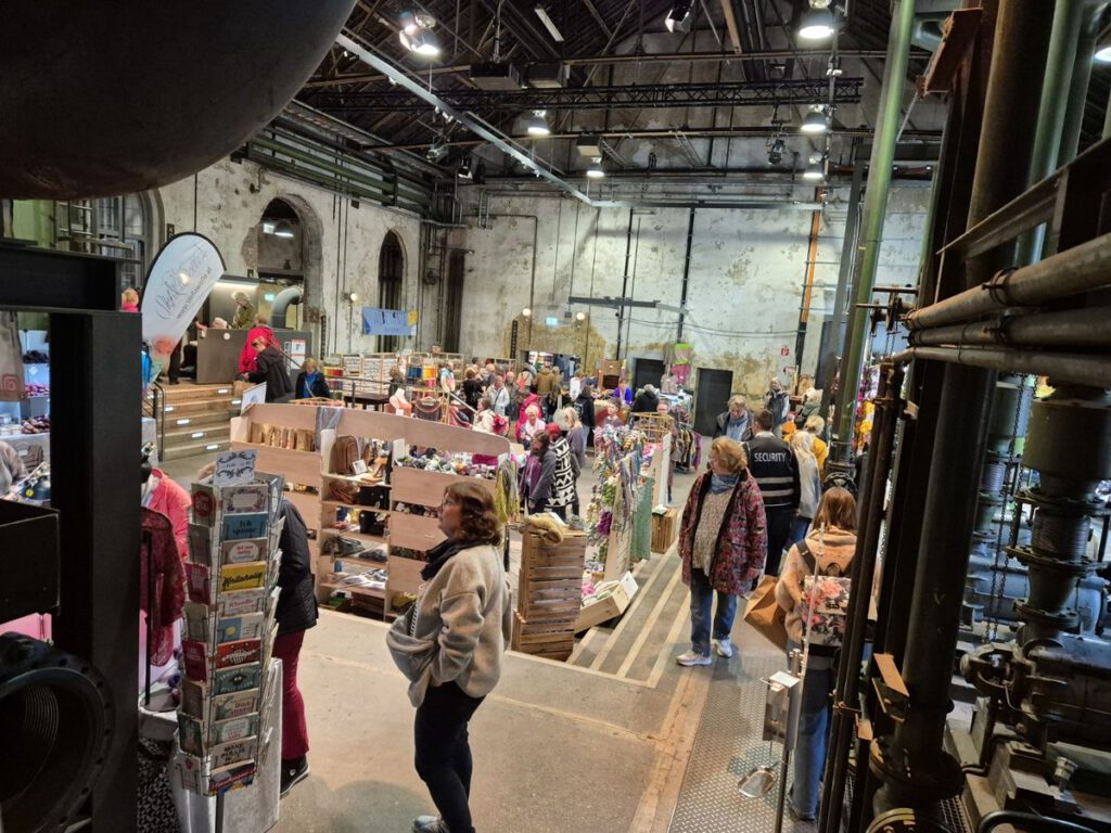 Blick in einen gut besuchten, engen Gang einer Messehalle auf dem Wollfest Kielgeholt mit vielen Menschen und bunten Wollständen